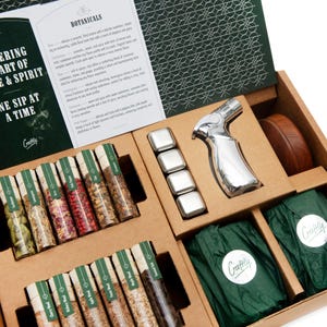 May include: A brown box with a green lid containing a set of small glass vials filled with various spices and herbs. The box also includes a silver torch, a wooden coaster, and a set of metal ice cubes. The box is labeled "Crafty" and "Botanicals".