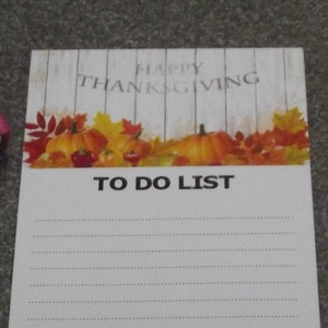 May include: A white notepad with a Thanksgiving theme. The top of the notepad has a woodgrain background with the text "Happy Thanksgiving" and a fall leaf design. The bottom of the notepad has the text "To Do List" and lined paper for writing.