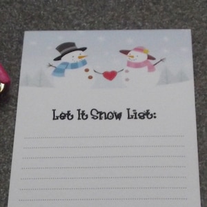 Pode incluir: Um bloco de notas branco com uma cena de inverno de dois bonecos de neve segurando um coração vermelho. O bloco de notas tem uma seção com linhas e o texto "Let It Snow List:" na parte superior.