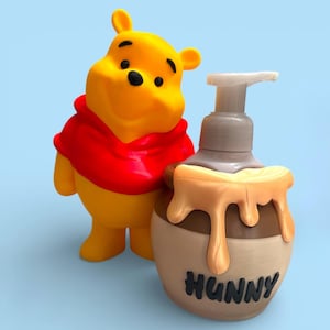 May include: A yellow Winnie the Pooh figurine stands next to a soap dispenser. The dispenser is shaped like a honey pot with the word "HUNNY" on it. The honey pot has a brown base with dripping honey accents.