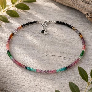 Könnte beinhalten: Eine Perlenkette mit kleinen, mehrfarbigen Edelsteinrondellen in Rosa-, Grün-, Blau- und Brauntönen, die in schwarze Perlen übergehen. Die Kette verfügt über einen silberfarbenen Karabinerverschluss, eine Verlängerungskette und einen kleinen Anhänger.