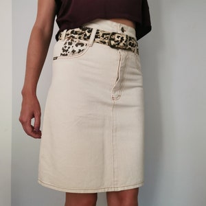 Könnte beinhalten: Ein cremefarbener Jeansrock mit einem Leopardenmuster-Gürtel. Der Rock hat eine gerade Silhouette und einen leicht ausgestellten Saum.