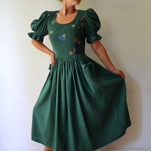 Puede incluir: Un vestido midi de color verde bosque con mangas cortas abullonadas con puños con volantes, escote redondo y corpiño ajustado. El vestido está adornado con pequeños motivos florales bordados en blanco, naranja y azul, e incluye dos bolsillos laterales.