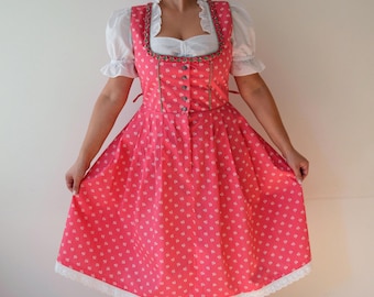 May include: A pink dirndl dress with a white heart pattern. The dress features a square neckline, a button-down bodice, and a full skirt. White lace trim adorns the hem. The dress is paired with a white blouse with ruffled sleeves.