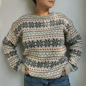 May include: A white, gray, and red patterned sweater with a crew neck. The sweater has a fair isle design with snowflakes and geometric patterns.