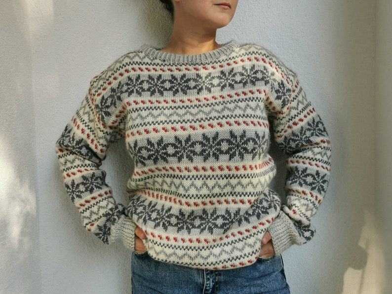 May include: A white, gray, and red patterned sweater with a crew neck. The sweater has a fair isle design with snowflakes and geometric patterns.