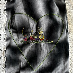May include: A black and white striped fabric with a green embroidered heart. Inside the heart, there are five frogs playing musical instruments. The fabric has a white lace trim.