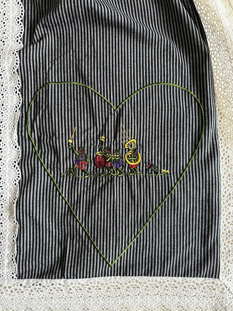May include: A black and white striped fabric with a green embroidered heart. Inside the heart, there are five frogs playing musical instruments. The fabric has a white lace trim.