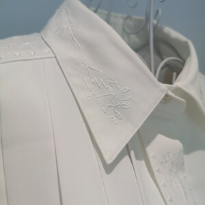 May include: White collared shirt with a delicate floral embroidery design on the collar and front. The shirt has a pleated front and a classic button-up closure.