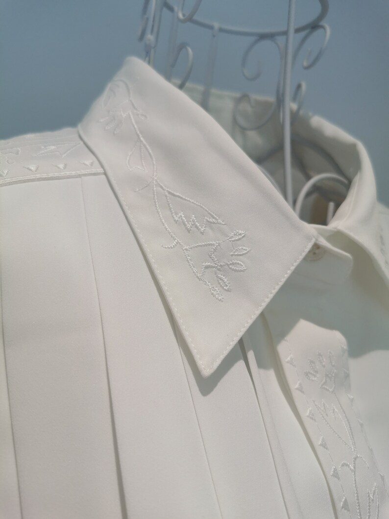 May include: White collared shirt with a delicate floral embroidery design on the collar and front. The shirt has a pleated front and a classic button-up closure.