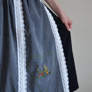 May include: A blue and white striped apron with white lace trim and an embroidered heart design. The heart features a colorful scene of people playing musical instruments.