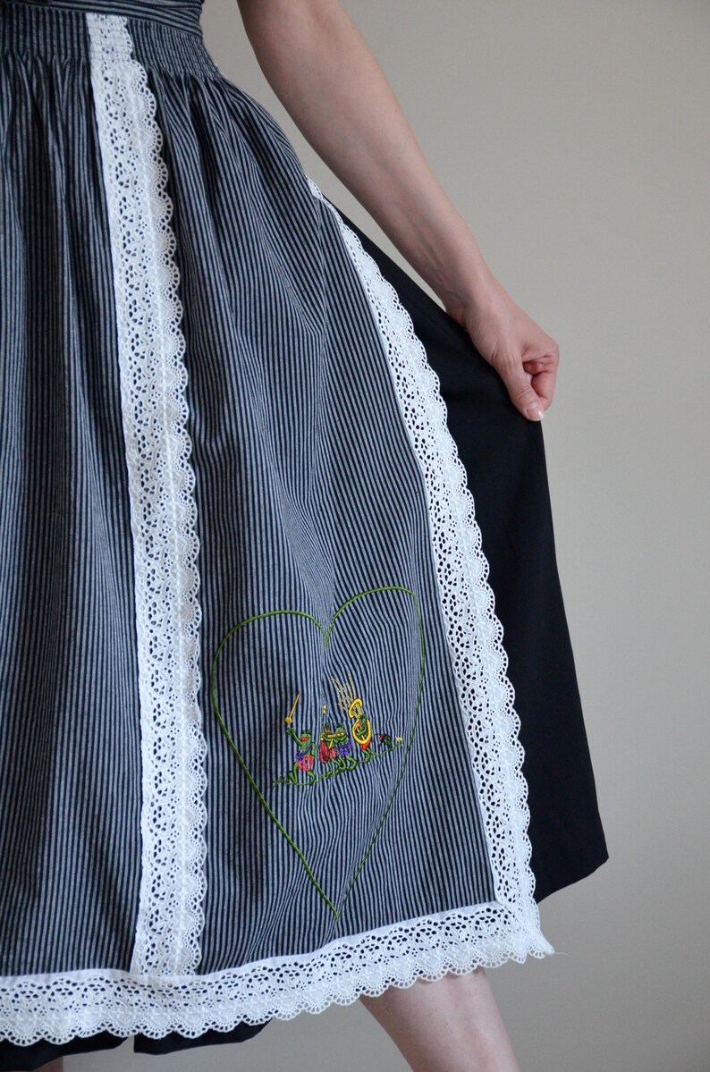 May include: A blue and white striped apron with white lace trim and an embroidered heart design. The heart features a colorful scene of people playing musical instruments.