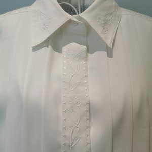 May include: White button-down shirt with a floral embroidery design on the front. The embroidery features a vine with delicate flowers.
