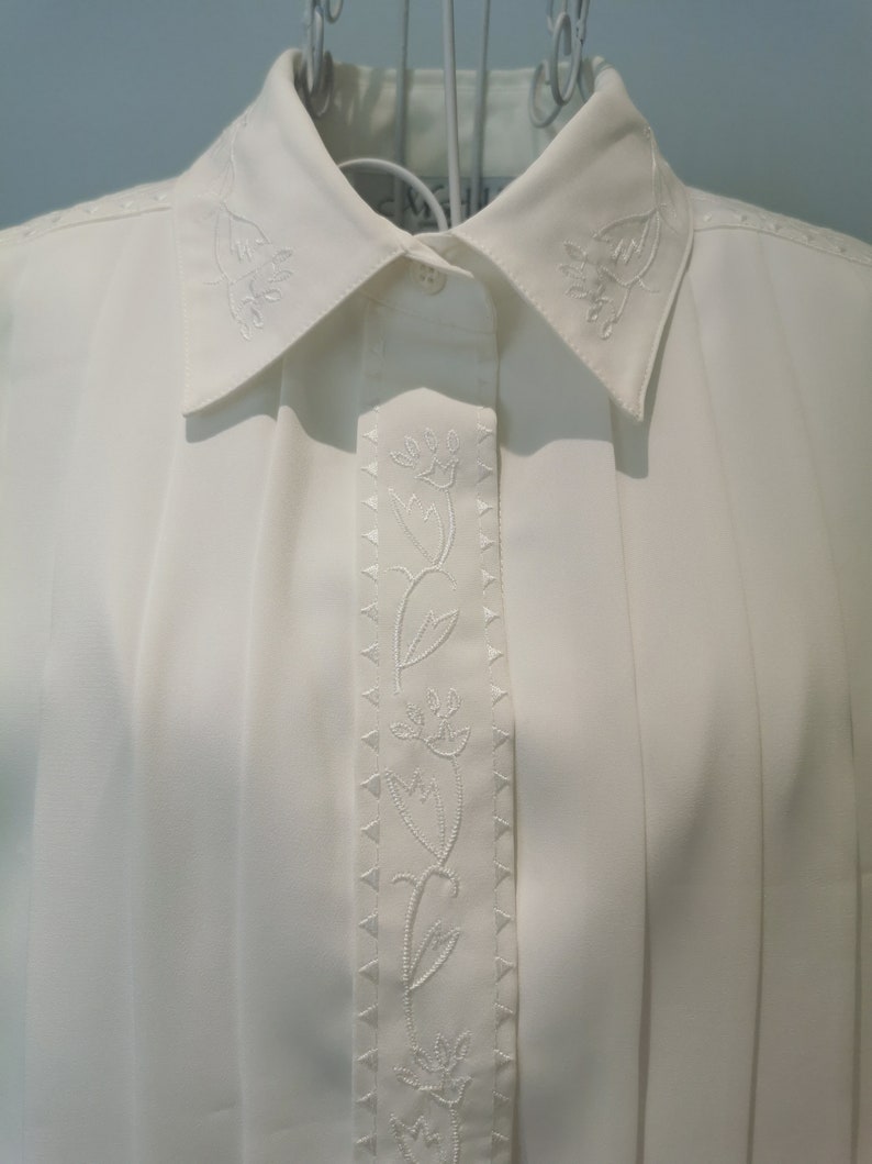 May include: White button-down shirt with a floral embroidery design on the front. The embroidery features a vine with delicate flowers.