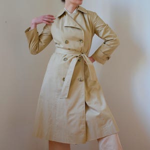 Op de afbeelding: Een beige trenchcoat met een dubbele rij knopen, een riem en een lange, vloeiende silhouet.
