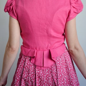 May include: A pink linen dirndl with a white floral print skirt and a ruffled hem. The bodice has a small embroidered logo on the back.