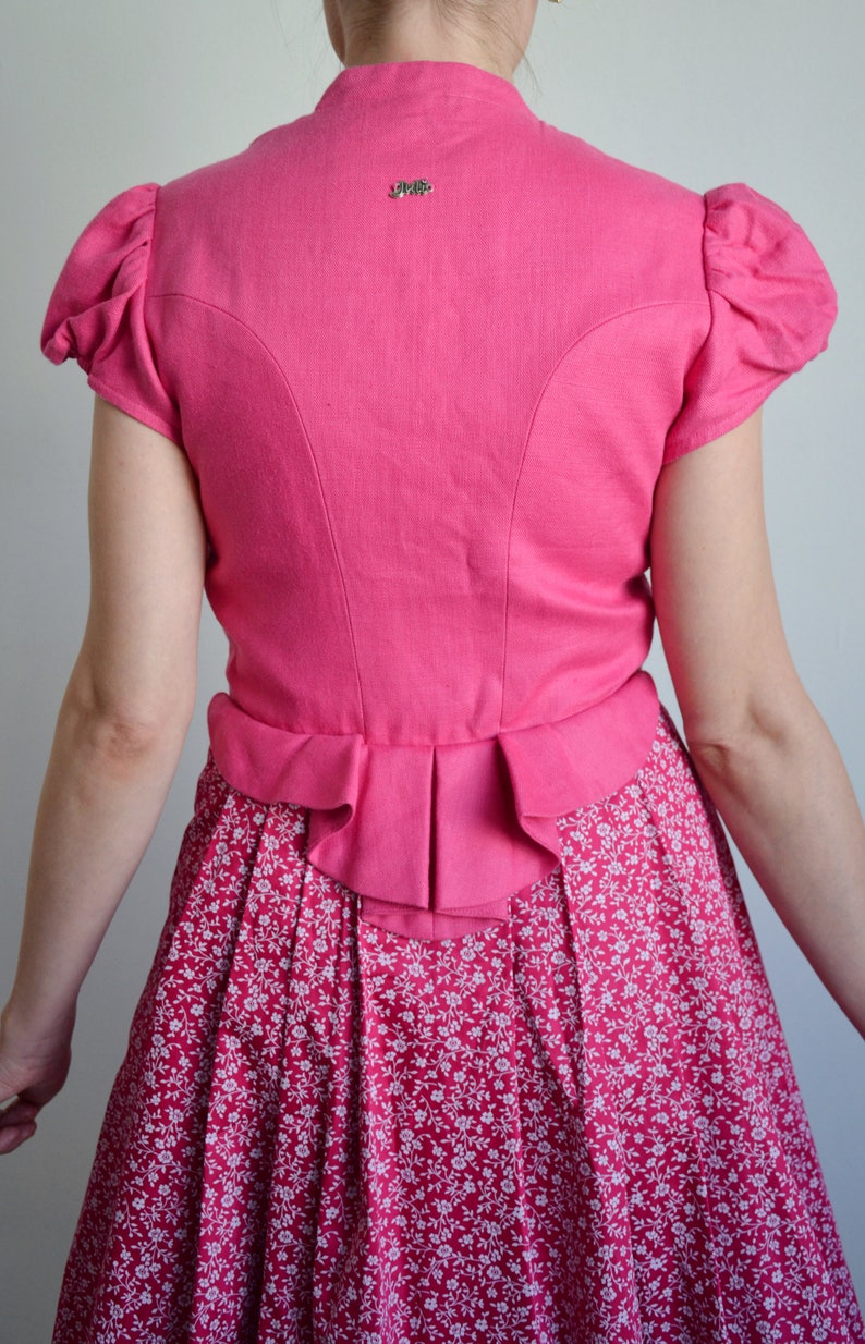 May include: A pink linen dirndl with a white floral print skirt and a ruffled hem. The bodice has a small embroidered logo on the back.