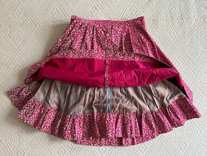 May include: A pink and white floral skirt with a ruffled hem and a button closure. The skirt has a lining and is made of a lightweight fabric.