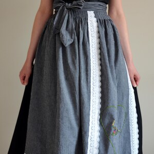 May include: A gray and white striped dirndl skirt with a white lace trim and a heart-shaped embroidered design. The skirt has a black ribbon tied at the waist.