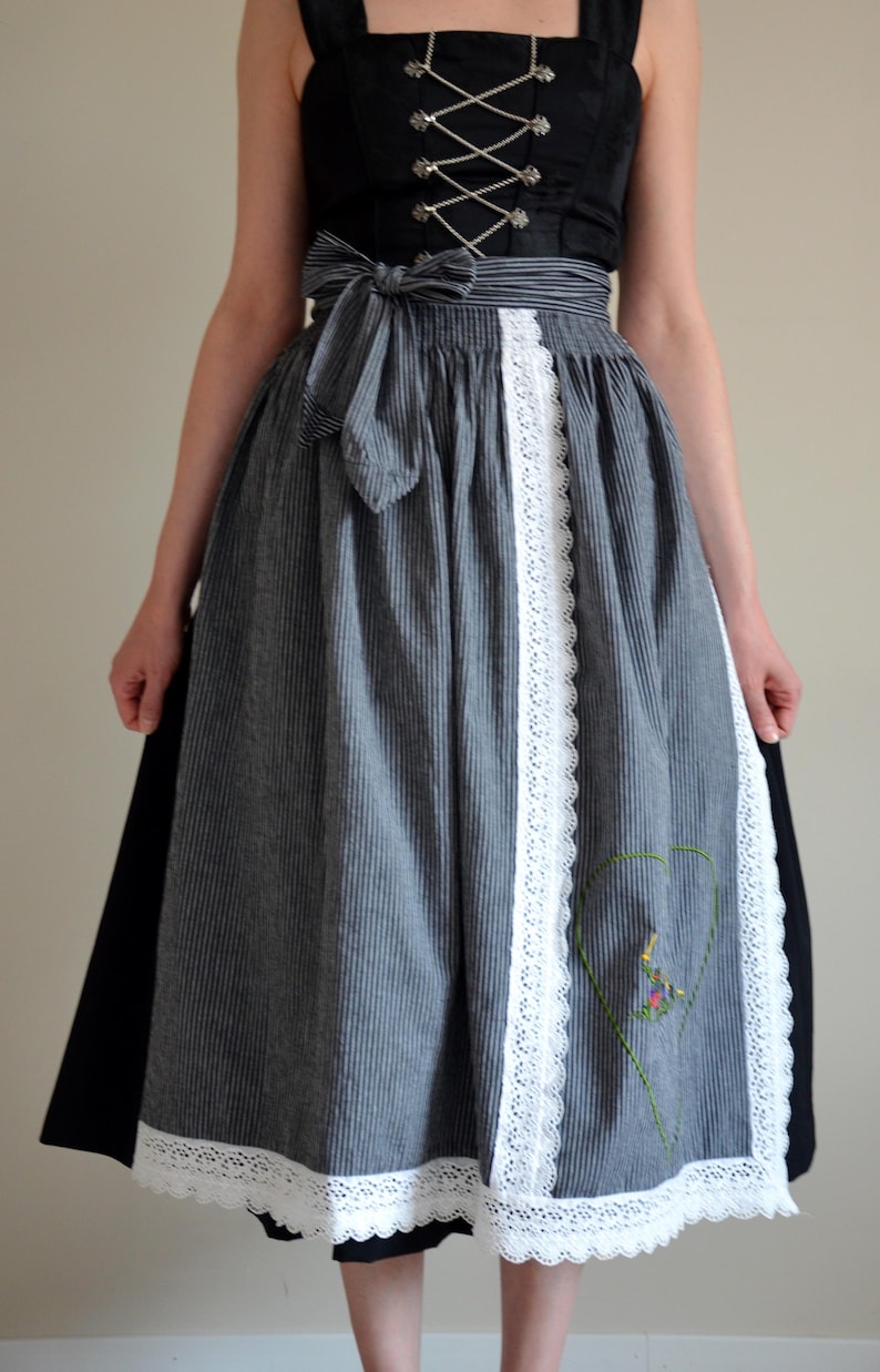 May include: A gray and white striped dirndl skirt with a white lace trim and a heart-shaped embroidered design. The skirt has a black ribbon tied at the waist.