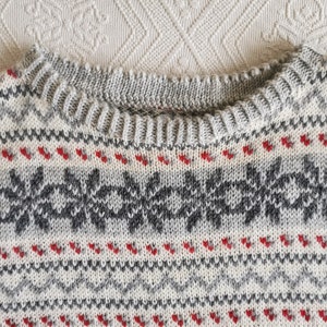 May include: Close-up of a grey and white knitted sweater with a red and grey snowflake pattern.