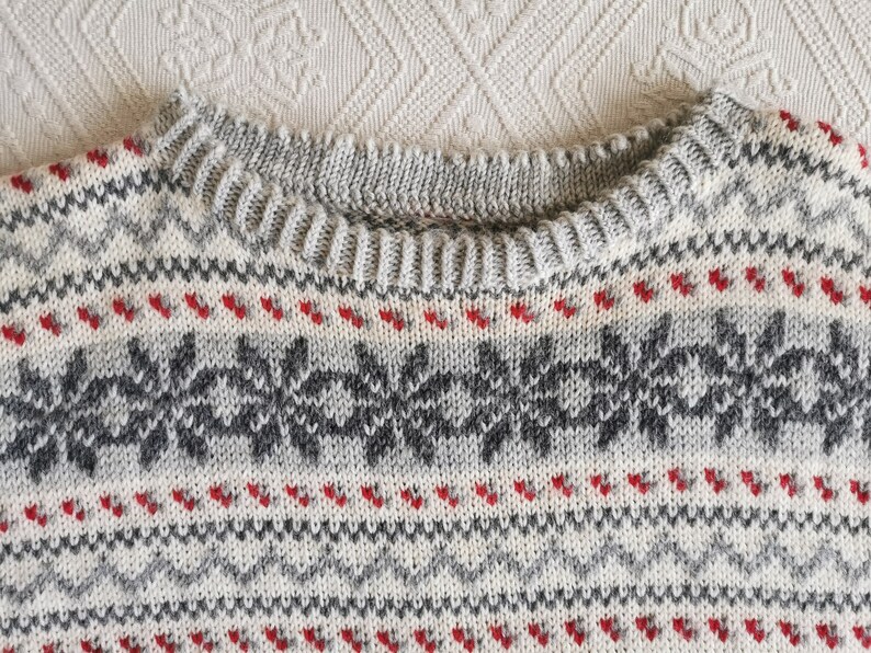May include: Close-up of a grey and white knitted sweater with a red and grey snowflake pattern.
