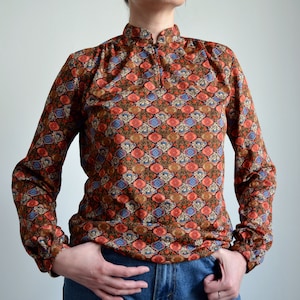 Op de afbeelding: Een lang mouw shirt met een bruin, oranje, blauw en wit geometrisch patroon. Het shirt heeft een hoge kraag en een ritssluiting.
