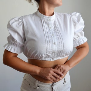 May include: White cropped blouse with a high collar, button-down front, and short, ruffled sleeves. The blouse has a fitted bodice and is made of a crisp, lightweight fabric. A classic, feminine top.