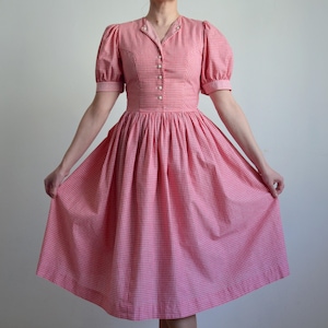 May include: A vintage-style, red and white gingham dress. The dress features a fitted bodice with a button-down front, short puffed sleeves, and a flared skirt. The dress is knee-length and has a classic, feminine silhouette.