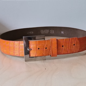 May include: Orange leather belt with a silver buckle. The belt has a crocodile pattern and the buckle has the text "REAL LEATHER" and "95" on it.
