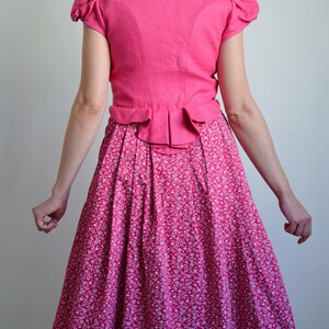 May include: A pink dirndl with a white floral print skirt and a matching pink bodice with a ruffled hem. The bodice has a short, puffed sleeve.