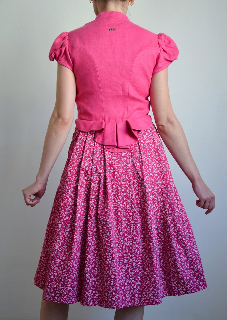 May include: A pink dirndl with a white floral print skirt and a matching pink bodice with a ruffled hem. The bodice has a short, puffed sleeve.