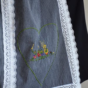 May include: A black and white striped apron with a white lace trim. The apron has a green heart-shaped design with an embroidered scene of frogs playing music.