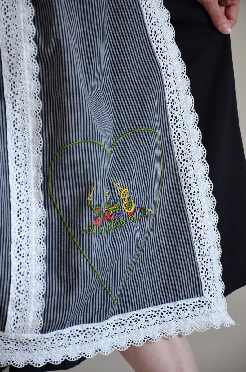 May include: A black and white striped apron with a white lace trim. The apron has a green heart-shaped design with an embroidered scene of frogs playing music.