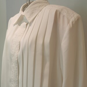 May include: A white long-sleeved blouse with a pleated front and embroidered floral details. The blouse has a classic collar and button closure.