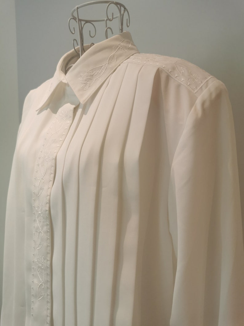 May include: A white long-sleeved blouse with a pleated front and embroidered floral details. The blouse has a classic collar and button closure.