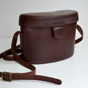 May include: A brown leather camera bag with a rounded top and a shoulder strap. The bag has a rectangular shape with a curved top and a buckle closure. The strap is adjustable and has a brass buckle.