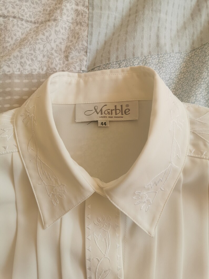 May include: White collared blouse with embroidered floral details. The blouse has a label that reads "Marble Ladies High Fashion" and "44".