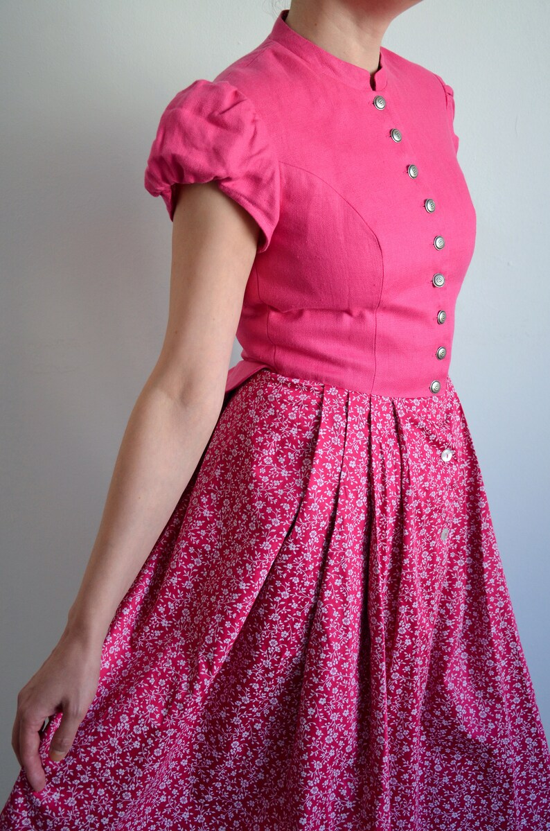 May include: A pink dress with a white floral print. The dress has a fitted bodice with a high neckline and short puffed sleeves. The skirt is full and gathered at the waist.