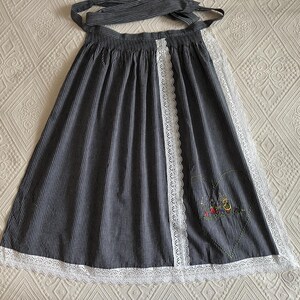 May include: A black and white striped apron with a white lace trim and a floral embroidery design. The apron has a tie at the waist and a long skirt.
