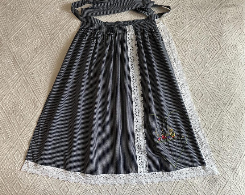 May include: A black and white striped apron with a white lace trim and a floral embroidery design. The apron has a tie at the waist and a long skirt.