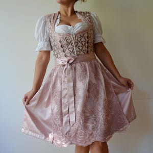 Könnte beinhalten: Ein traditionelles rosa Dirndlkleid mit weißer Bluse und dekorativem Mieder. Das Kleid hat einen Satinstock mit Spitzendetails und einen passenden Schleifengürtel. Das Mieder hat ein verziertes Muster.