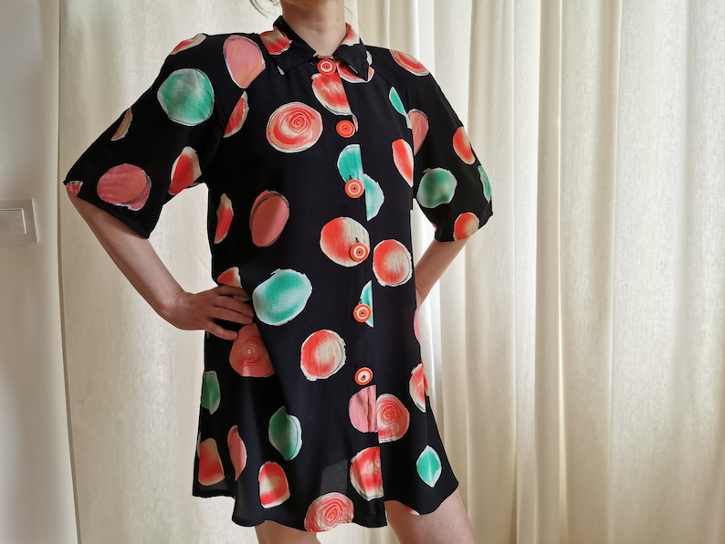 May include: A black short-sleeved button-up dress with a colorful abstract print of large circles in shades of red, pink, and green. The dress has a collared neckline and a button-up front.