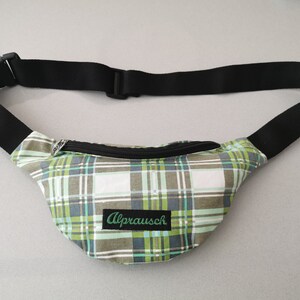May include: A green, white, and black plaid fanny pack with a black adjustable strap and a zipper closure. The fanny pack has the word "Alprausch" embroidered on the front.