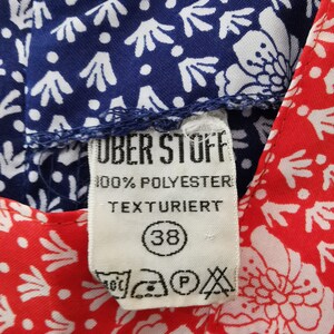 May include: A white tag with blue text that reads "Uber Stoff 100% Polyester Texturiert" and a black circle with the number 38 inside. The tag is attached to a blue and white patterned fabric and a red and white patterned fabric.