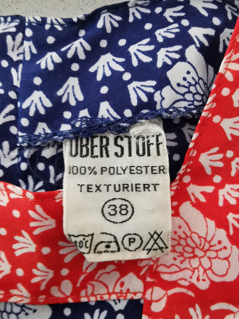 May include: A white tag with blue text that reads "Uber Stoff 100% Polyester Texturiert" and a black circle with the number 38 inside. The tag is attached to a blue and white patterned fabric and a red and white patterned fabric.