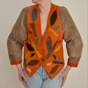 May include: An orange suede jacket with brown sleeves and colorful leaf-like appliqués. The jacket has a V-neck design and is worn over blue jeans. The sleeves are a contrasting brown color with orange trim.