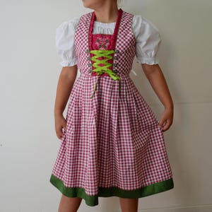 Peut inclure: Une robe dirndl traditionnelle à carreaux rouges et blancs. La robe présente un corsage avec des lacets verts, un accent brodé floral et un chemisier blanc à manches bouffantes. La jupe a un ourlet vert.