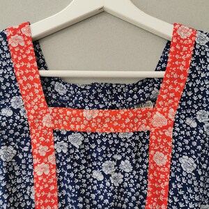 May include: A blue and white floral print dress with red trim around the neckline and shoulders. The dress has a square neckline and is hanging on a white hanger. The label "UBER STORY" is visible on the neckline.