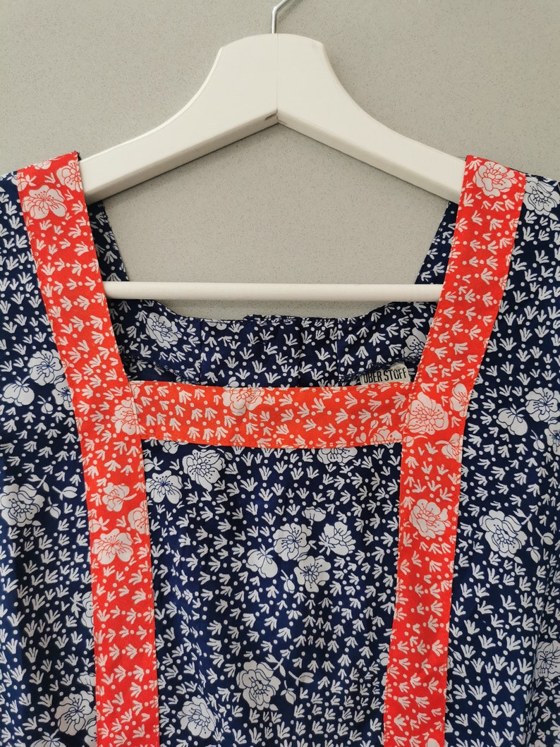 May include: A blue and white floral print dress with red trim around the neckline and shoulders. The dress has a square neckline and is hanging on a white hanger. The label "UBER STORY" is visible on the neckline.